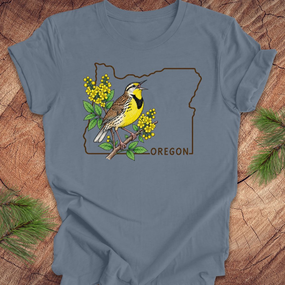 A slate blue short-sleeve t-shirt laid flat on a rustic wood background with pine branches in the corners. The shirt features a graphic of a yellow and black bird perched on a branch with yellow flowers, framed by the outline of Oregon and the word 'OREGON'.