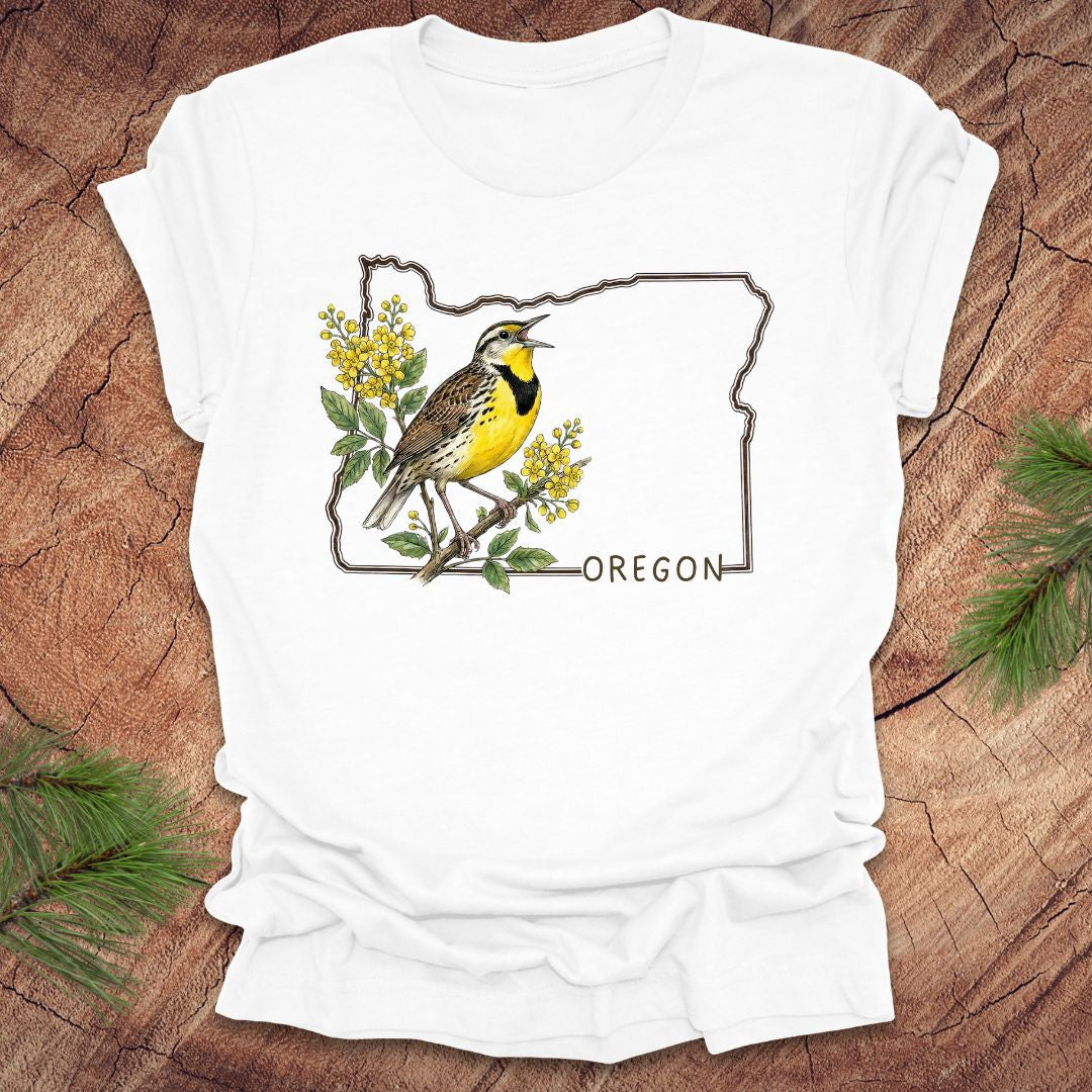 White t-shirt with a bird and floral design on an outline of Oregon, placed on a wooden surface.