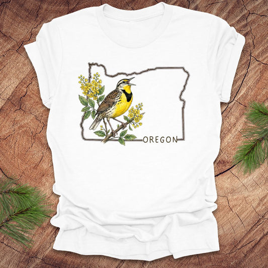 White t-shirt with a bird and floral design on an outline of Oregon, placed on a wooden surface.