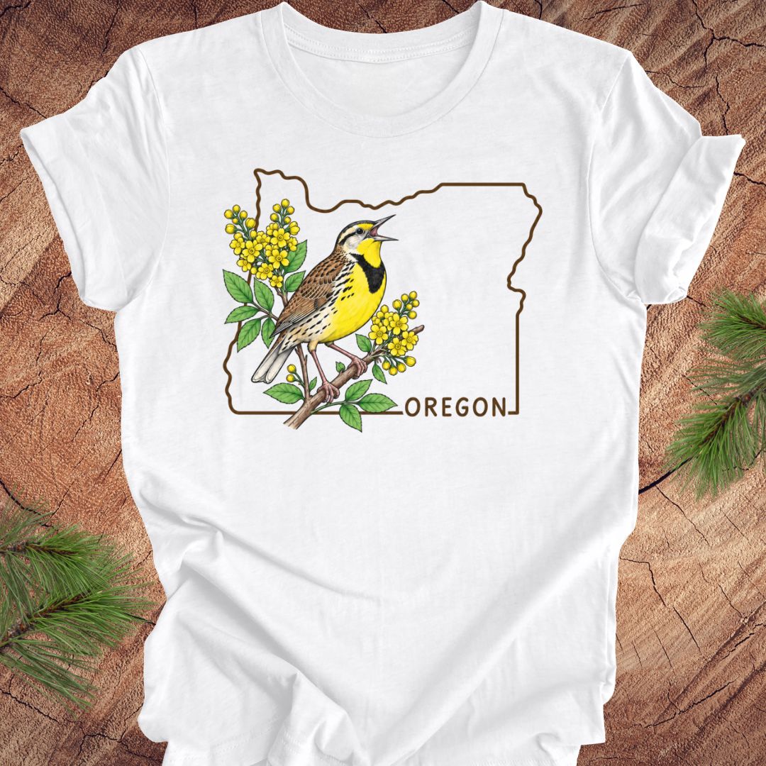 A white short-sleeve crewneck t-shirt laid flat on a rustic wood background with pine branches in the corners. The shirt features a graphic print of a yellow and brown bird perched on a branch with yellow flowers, framed by the outline of Oregon and the word 'OREGON' printed below.