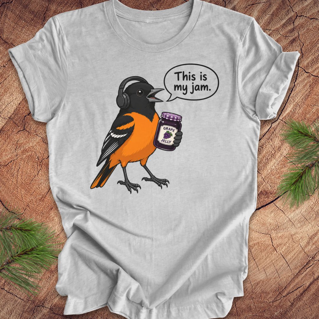 A light gray short-sleeve t-shirt laid flat on a rustic wood surface with pine branches in the corners. The shirt features a graphic of a Baltimore Oriole bird wearing headphones and holding a jar labeled 'GRAPE JAM', with a speech bubble that reads 'This is my jam.
