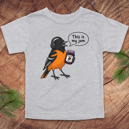 A light heather gray short-sleeve t-shirt with a cartoon graphic of a bird wearing headphones and holding a jar of jam, with a speech bubble that reads 'This is my jam.' The shirt is laid flat on a rustic wood background with pine branches in the corners.
