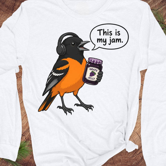 A white long-sleeve t-shirt with a graphic of an orange and black bird wearing headphones and holding a jar of grape jelly, with a speech bubble that reads 'This is my jam.' The shirt is laid flat on a rustic wood background.