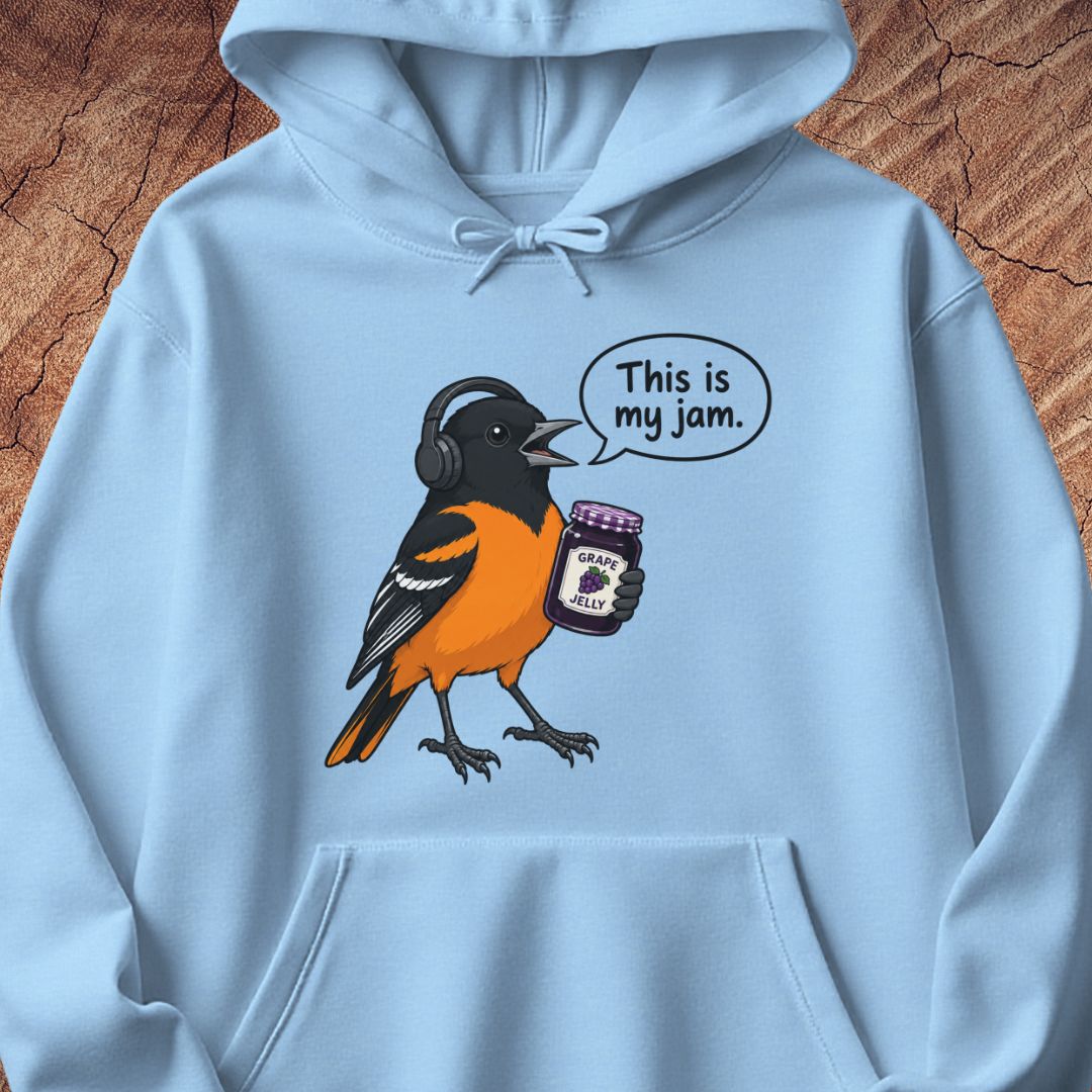 A light blue hoodie laid flat on a textured brown wood background, featuring a cartoon bird graphic wearing black headphones and holding a jar of jam, with a speech bubble that reads 'This is my jam.'.