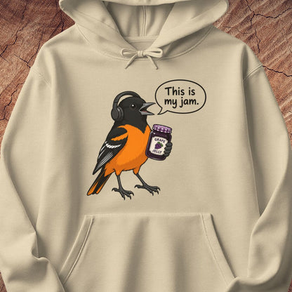 A beige hoodie featuring a cartoon bird wearing headphones and holding a jar of 'Grape Jelly' with a speech bubble that reads 'This is my jam.' The hoodie is displayed flat against a textured brown wood background.