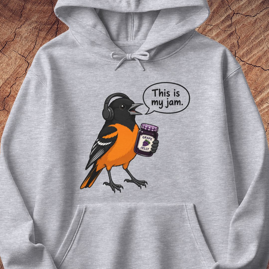 A light heather gray pullover hoodie featuring a cartoon graphic of an orange and black bird wearing headphones and holding a jar of jam, with a speech bubble that reads 'This is my jam.' The hoodie is displayed flat against a rustic wood background.