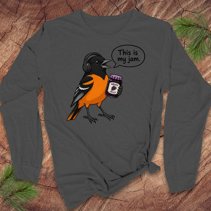 A dark gray long-sleeve t-shirt with a graphic of an oriole bird wearing headphones and holding a jar of jam, with a speech bubble that says 'This is my jam.' The shirt is displayed flat on a rustic wood background with pine branches in the corners.