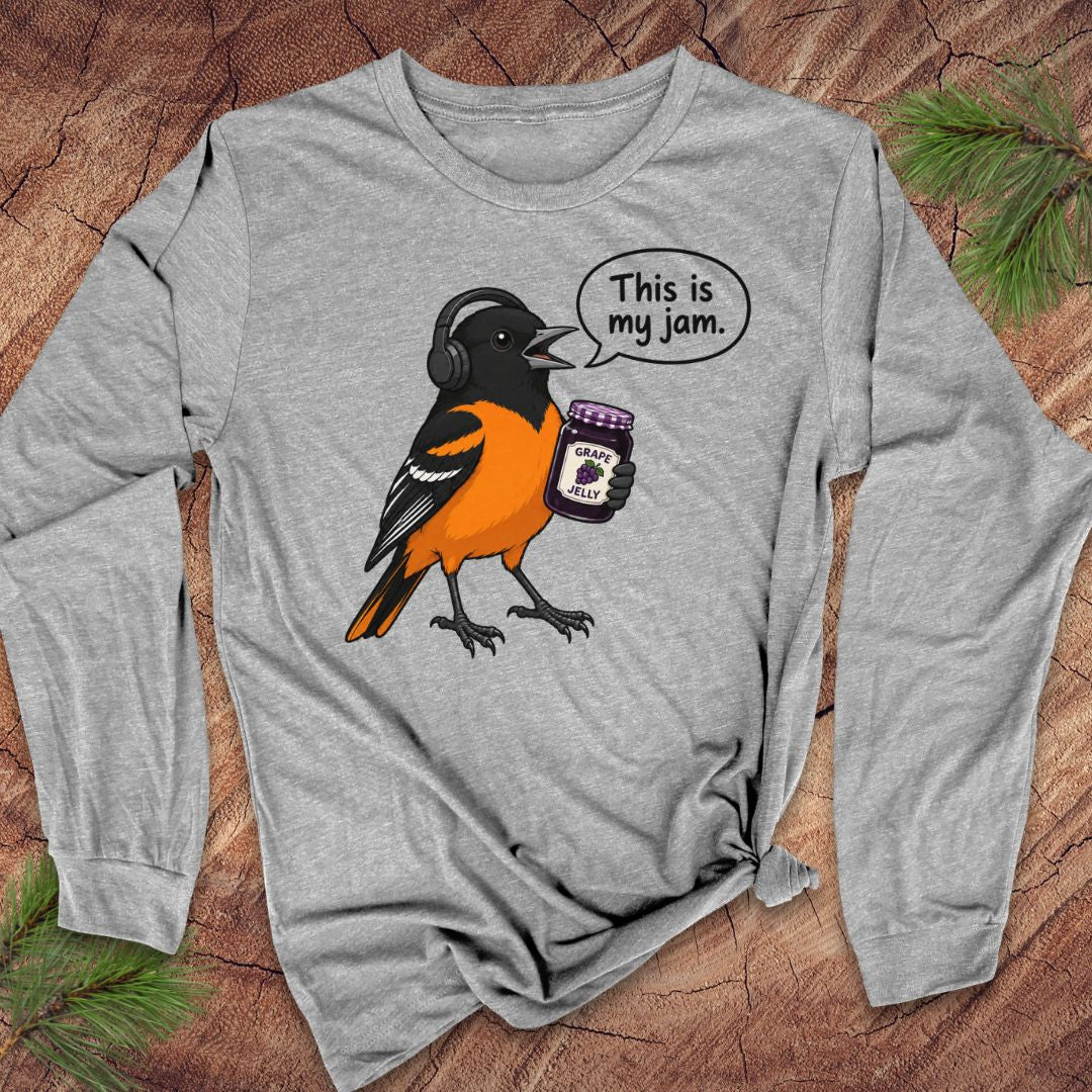 A heather gray long-sleeve t-shirt with a graphic of an oriole bird wearing headphones and holding a jar of jam, with a speech bubble that says 'This is my jam.' The shirt is laid flat on a rustic wood background with pine needles.