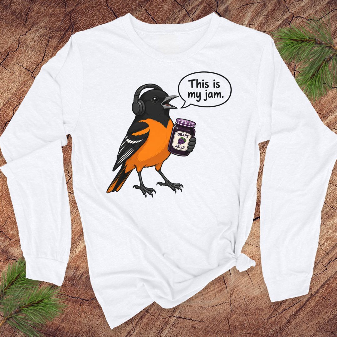 A white long-sleeve t-shirt with a graphic of an oriole bird wearing headphones and holding a jar of jam, with a speech bubble that says 'This is my jam.' The shirt is laid flat on a rustic wood background with pine branches in the corners.