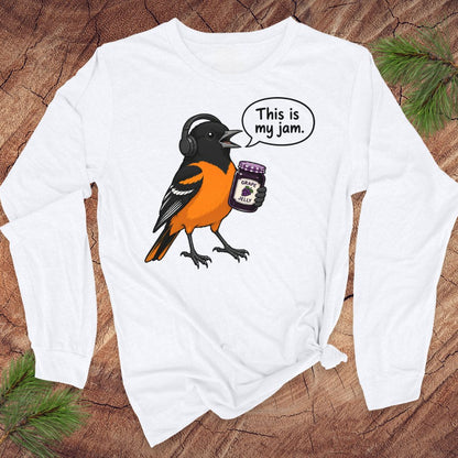 A white long-sleeve t-shirt with a graphic of an oriole bird wearing headphones and holding a jar of jam, with a speech bubble that says 'This is my jam.' The shirt is laid flat on a rustic wood background with pine branches in the corners.