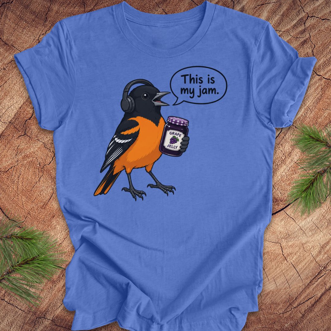 A blue short-sleeve crewneck t-shirt laid flat on a rustic wood background with pine needles in the corners. The shirt features a graphic of a Baltimore Oriole bird wearing headphones and holding a jar of grape jelly, with a speech bubble that reads 'This is my jam.