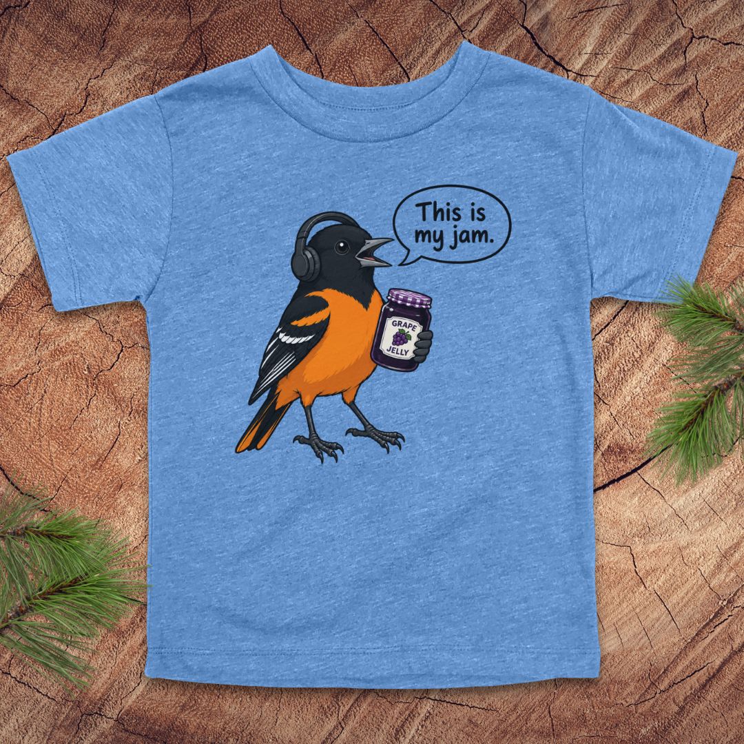 A heather blue short-sleeve t-shirt featuring a cartoon orishrike holding a jar of jam with a speech bubble that says 'This is my jam.' The shirt is laid flat on a rustic wood background with pine needles in the corners.