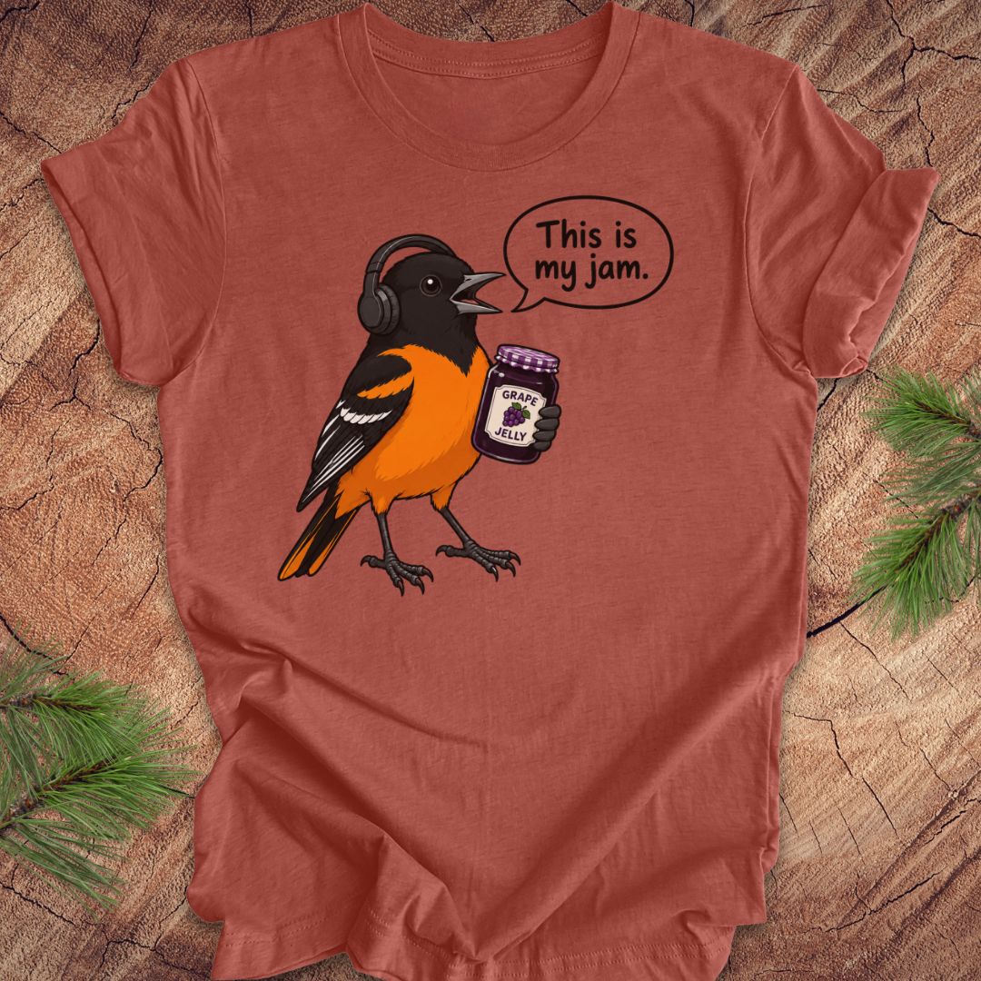 A terracotta-colored short-sleeve t-shirt laid flat on a rustic wood background with pine branches in the corners. The shirt features a cartoon graphic of a Baltimore Oriole bird wearing headphones and holding a jar of grape jelly, with a speech bubble that reads 'This is my jam.