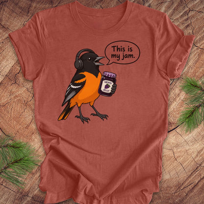 A terracotta-colored short-sleeve t-shirt laid flat on a rustic wood background with pine branches in the corners. The shirt features a cartoon graphic of a Baltimore Oriole bird wearing headphones and holding a jar of grape jelly, with a speech bubble that reads 'This is my jam.