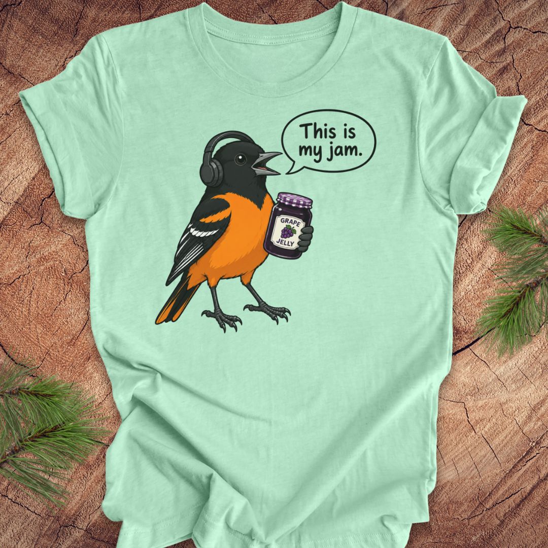 A mint green short-sleeve t-shirt laid flat on a rustic wood surface, featuring a cartoon Oriole bird wearing headphones and holding a jar of grain jelly with a speech bubble that says 'This is my jam.'.
