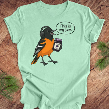 A mint green short-sleeve t-shirt laid flat on a rustic wood surface, featuring a cartoon Oriole bird wearing headphones and holding a jar of grain jelly with a speech bubble that says 'This is my jam.'.