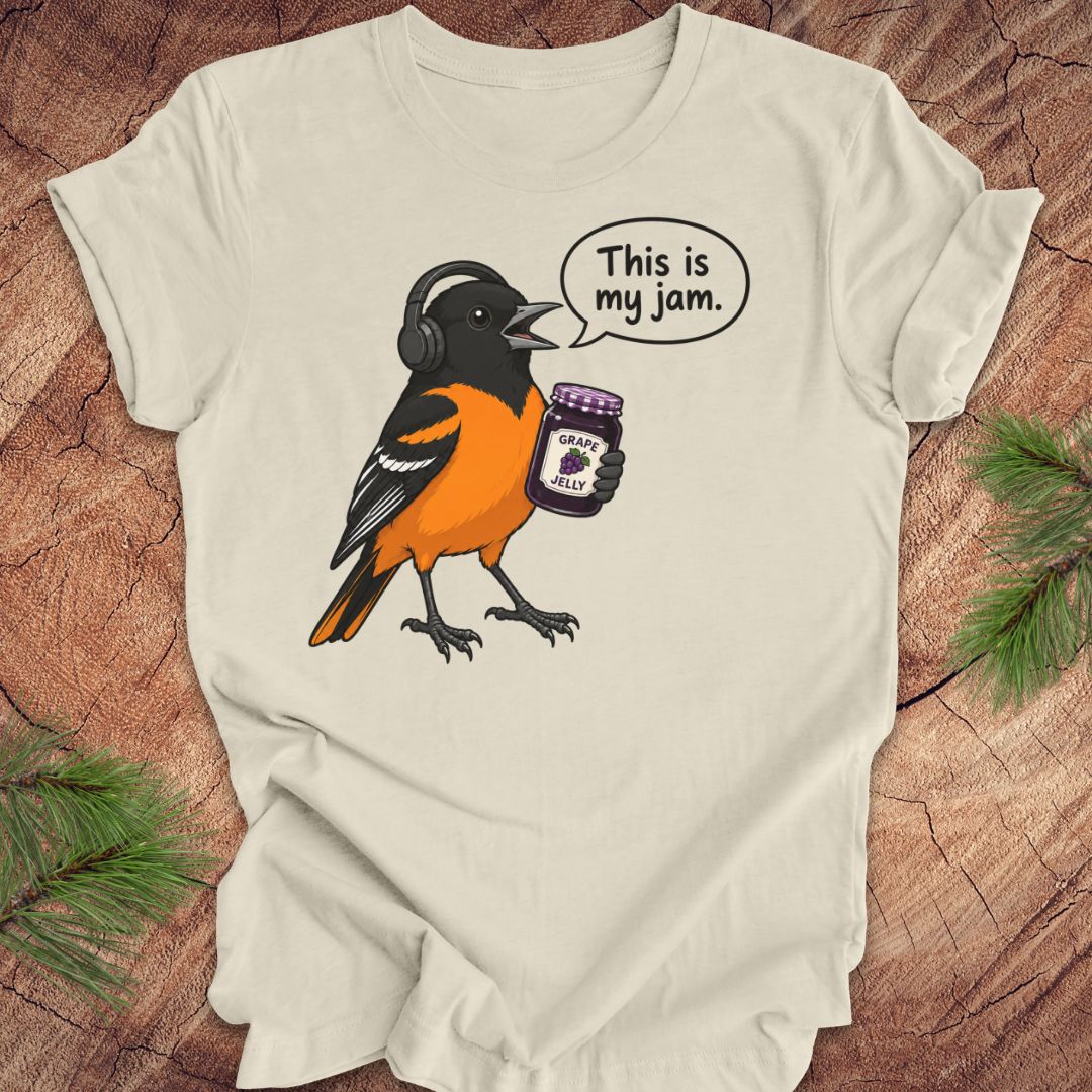A beige short-sleeve t-shirt laid flat on a rustic wood background, featuring a cartoon graphic of a Baltimore Oriole bird wearing headphones and holding a jar of grape jelly, with a speech bubble that reads 'This is my jam.'.