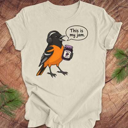 A beige short-sleeve t-shirt laid flat on a rustic wood background, featuring a cartoon graphic of a Baltimore Oriole bird wearing headphones and holding a jar of grape jelly, with a speech bubble that reads 'This is my jam.'.