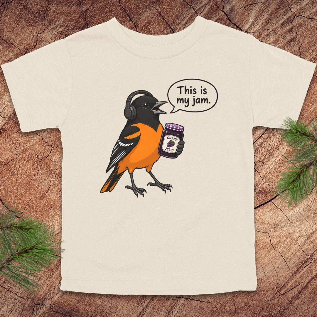 An off-white short-sleeve t-shirt laid flat on a rustic wood background with pine needles in the corners. The shirt features a cartoon graphic of a bird wearing black headphones and holding a jar of jam, with a speech bubble that reads 'This is my jam.