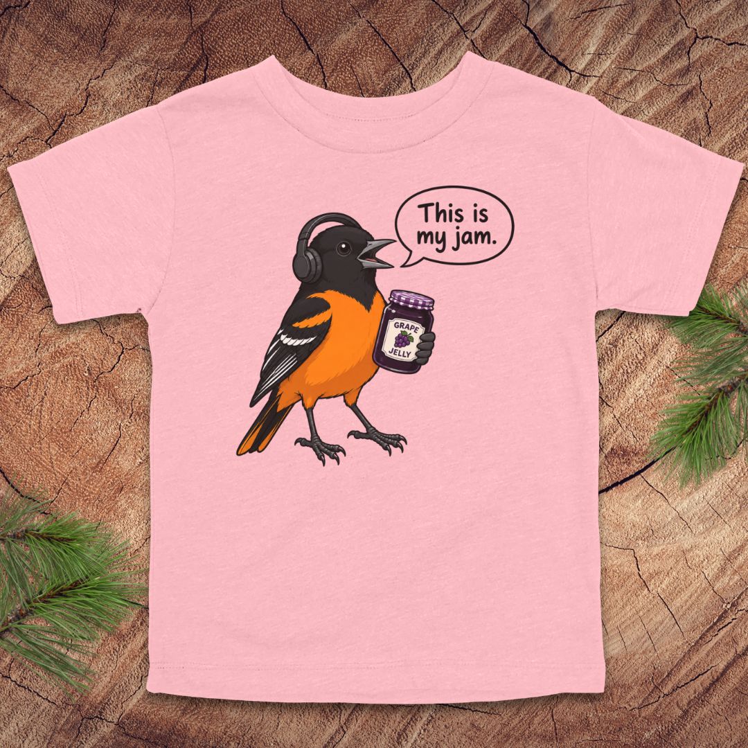 A light pink short-sleeve t-shirt laid flat on a rustic wood background with pine branches in the corners. The shirt features a cartoon illustration of a bird wearing headphones and holding a jar, with a speech bubble that reads 'This is my jam.