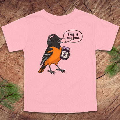 A light pink short-sleeve t-shirt laid flat on a rustic wood background with pine branches in the corners. The shirt features a cartoon illustration of a bird wearing headphones and holding a jar, with a speech bubble that reads 'This is my jam.