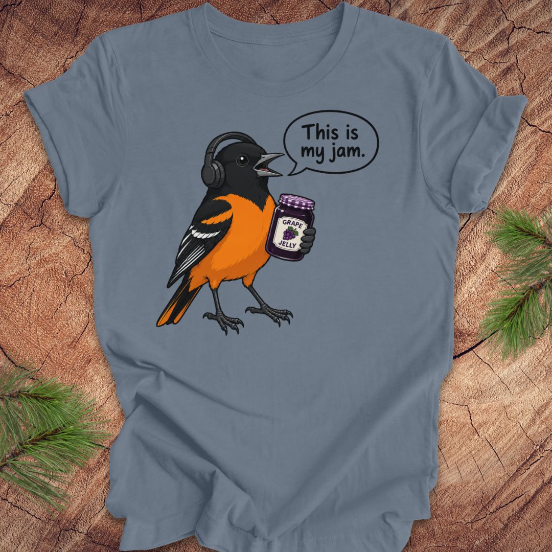 A slate blue short-sleeve t-shirt laid flat on a rustic wood background, featuring a cartoon graphic of an Oriole bird wearing headphones and holding a jar of grape jelly, with a speech bubble that reads 'This is my jam.'.