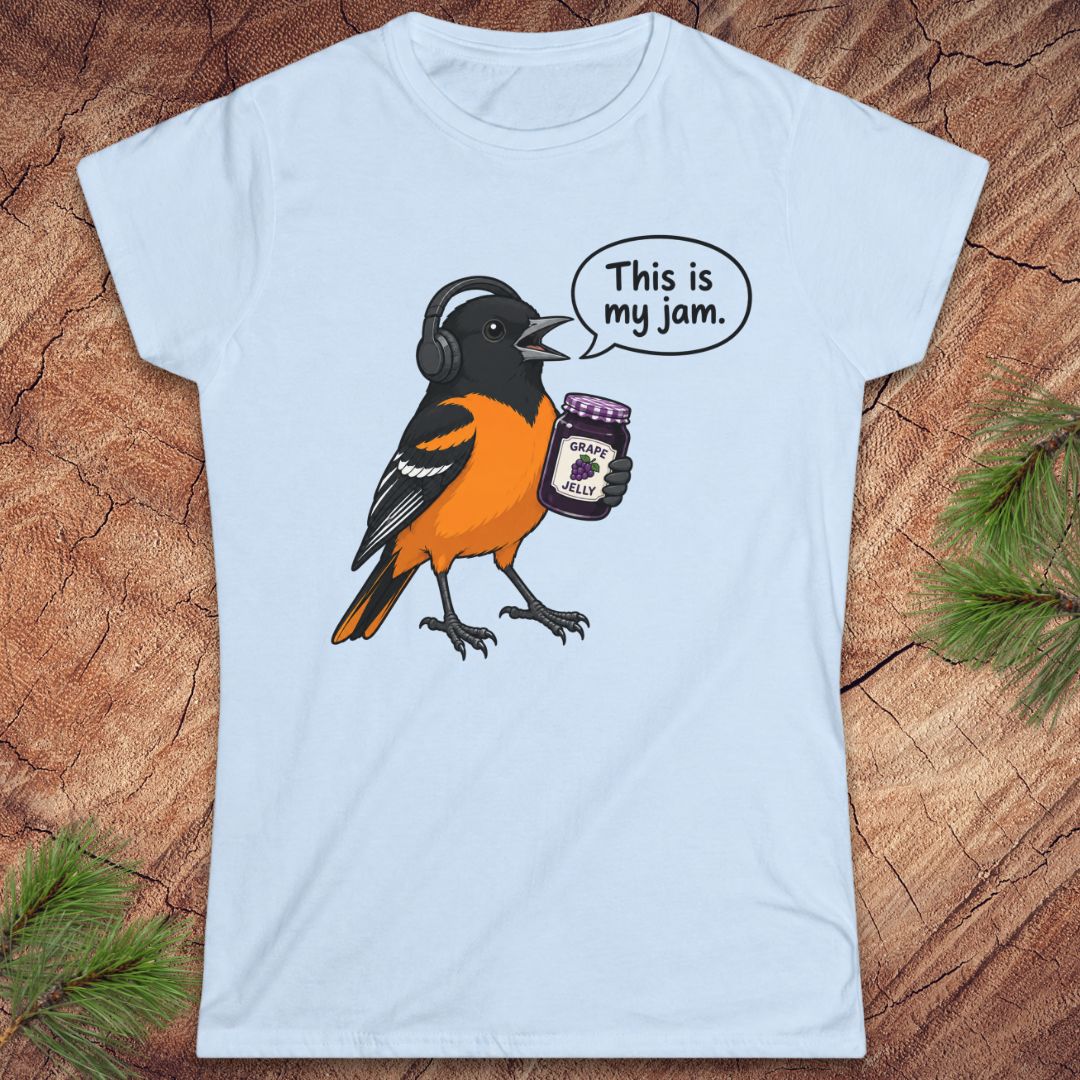 A light blue women's short-sleeve t-shirt laid flat on a rustic wood background with pine needles. The shirt features a cartoon graphic of a Baltimore Oriole bird wearing headphones and holding a jar of 'Peanut Butter Jams', with a speech bubble that reads 'This is my jam.