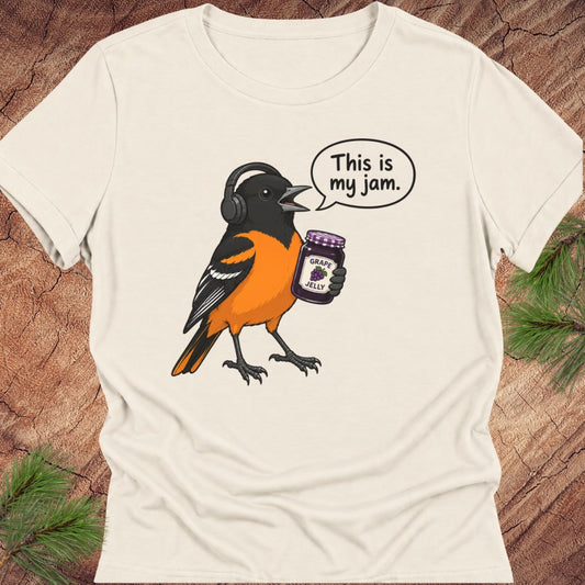 Natural cream women's relaxed t-shirt with a Baltimore Oriole in headphones holding a grape jelly jar and a 'This is my jam.' speech bubble, laid flat on a rustic wood background with pine branches.