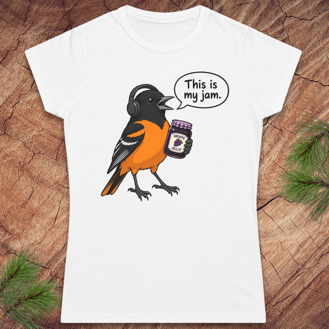 A white short-sleeve crewneck t-shirt featuring a cartoon graphic of a Baltimore Oriole bird wearing headphones and holding a jar of 'Peanut Butter Jams', with a speech bubble that reads 'This is my jam.' The shirt is displayed flat on a rustic wood background with pine needles in the corners.