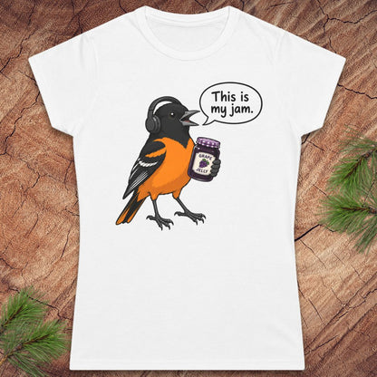 A white short-sleeve crewneck t-shirt featuring a cartoon graphic of a Baltimore Oriole bird wearing headphones and holding a jar of 'Peanut Butter Jams', with a speech bubble that reads 'This is my jam.' The shirt is displayed flat on a rustic wood background with pine needles in the corners.