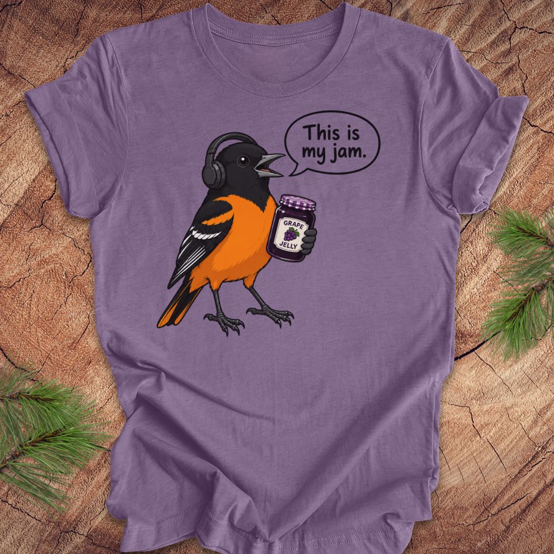 A purple short-sleeve t-shirt laid flat on a rustic wood surface, featuring a cartoon Oriole bird wearing black headphones and holding a jar of grape jelly, with a speech bubble that reads 'This is my jam.'.