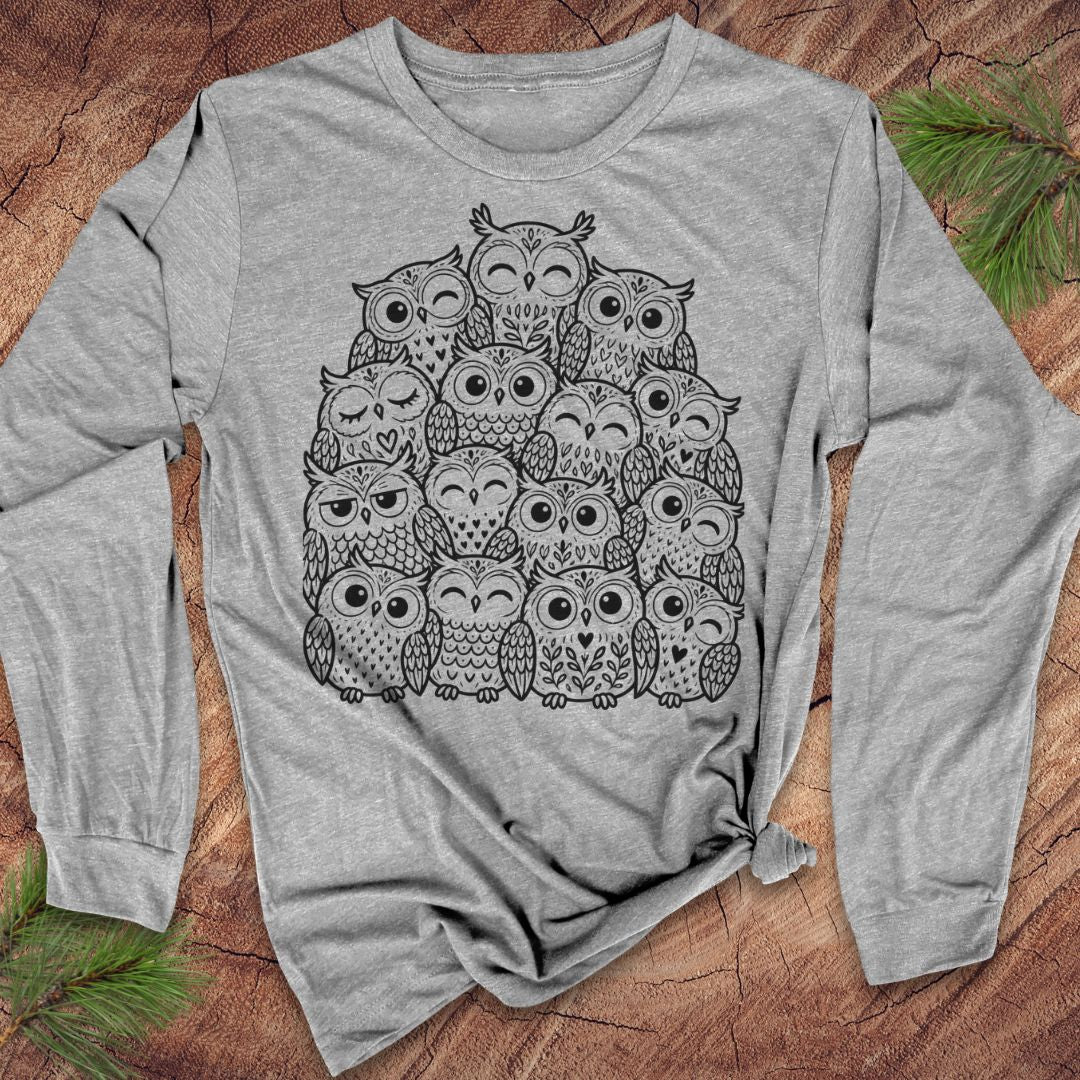 Athletic heather gray long-sleeve t-shirt with a black line-art huddle of cute owls piled together, laid flat on a rustic wood background with pine branches.