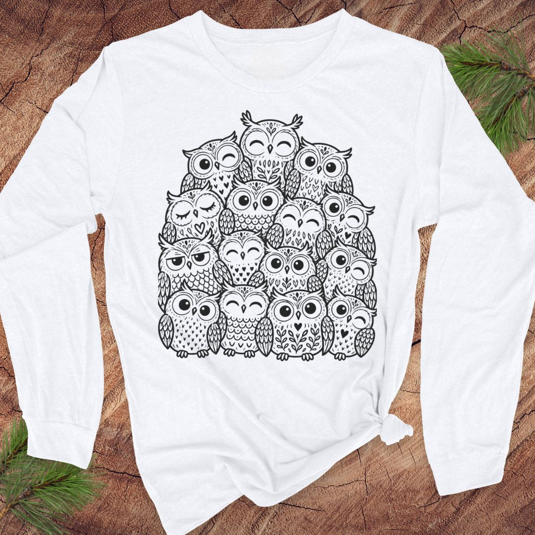 White long-sleeve t-shirt with a black line-art huddle of cute owls piled together, laid flat on a rustic wood background with pine branches.