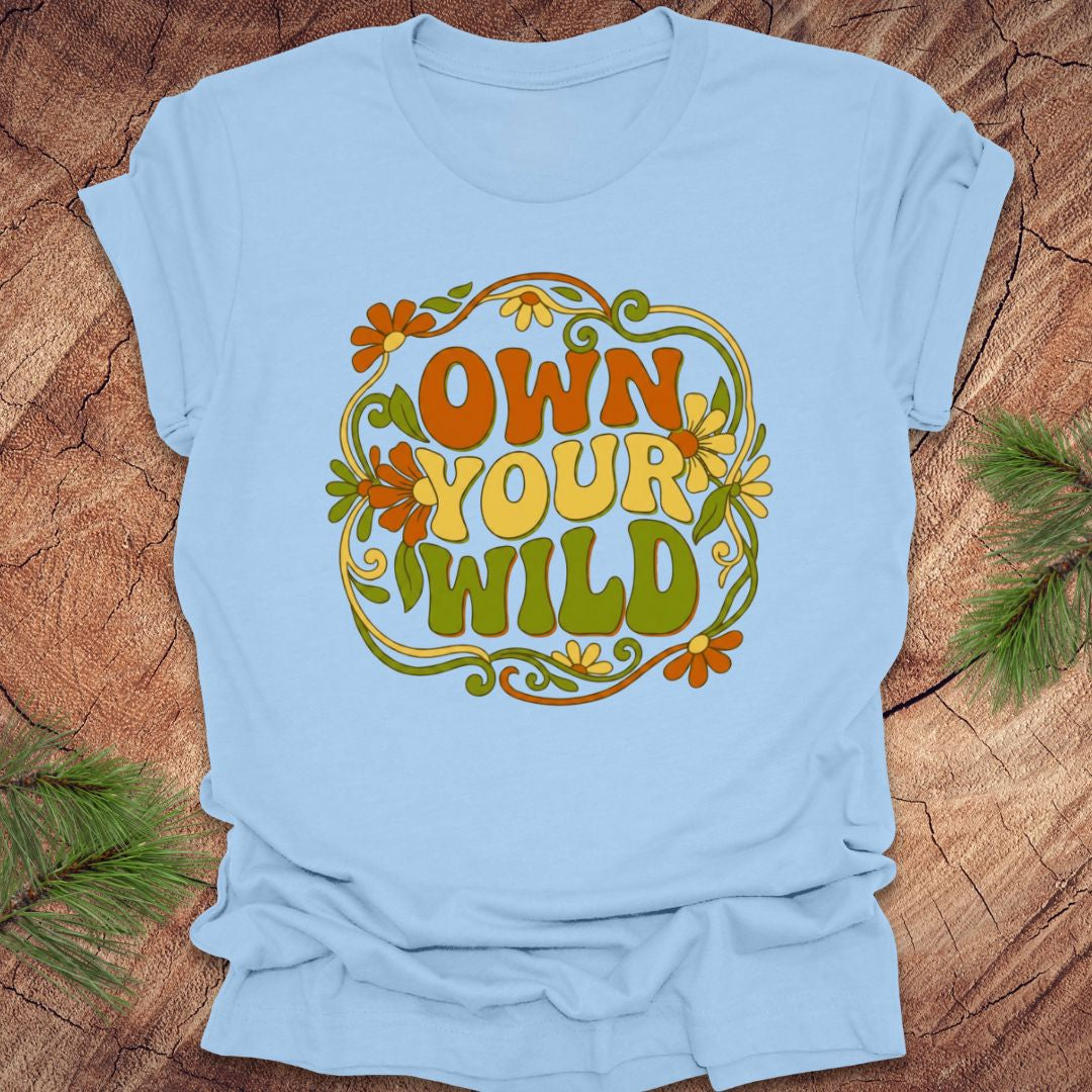 Light blue t-shirt with 'Own Your Wild' floral design on a wooden surface with greenery.