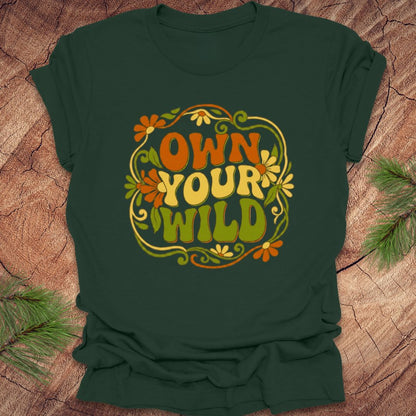 Green t-shirt with 'Own Your Wild' floral design on a wooden surface with greenery.