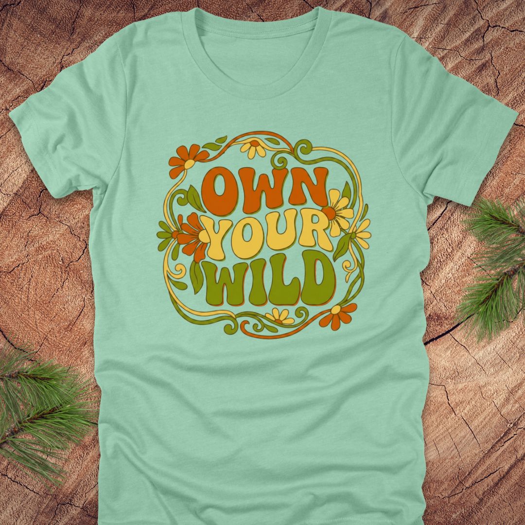 Mint green t-shirt with 'Own Your Wild' floral design on a wooden surface with greenery.