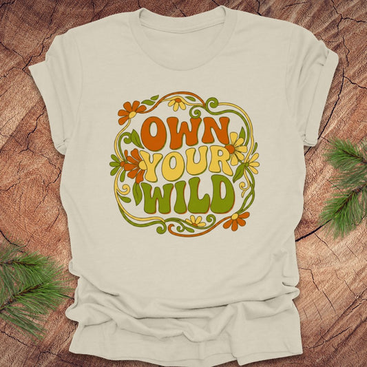 Beige t-shirt with 'Own Your Wild' floral design on a wooden surface with greenery.