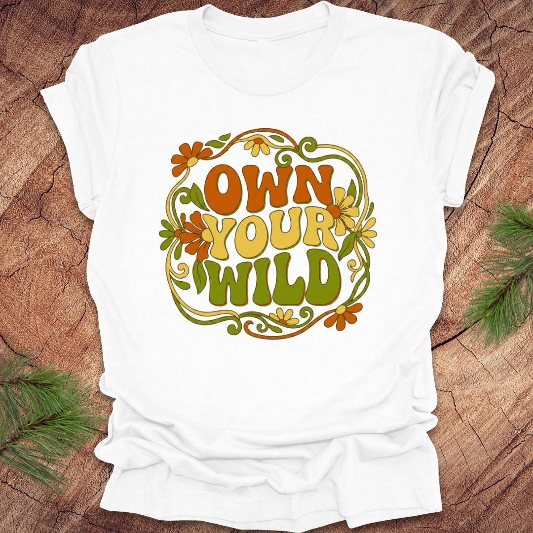 White t-shirt with 'Own Your Wild' floral design on a wooden surface with greenery.