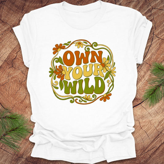 White t-shirt with 'Own Your Wild' floral design on a wooden surface with greenery.