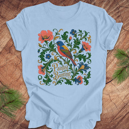 Blue t-shirt with a painted bunting bird and floral design on a wood background.