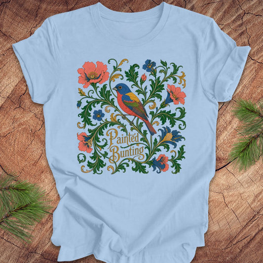 Blue t-shirt with a painted bunting bird and floral design on a wood background.