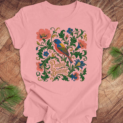 Pink t-shirt with a painted bunting bird and floral design on a wood background.