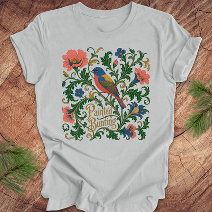 Gray t-shirt with a painted bunting bird and floral design on a wood background.