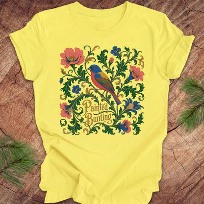 Yellow t-shirt with a painted bunting bird and floral design on a wood background.