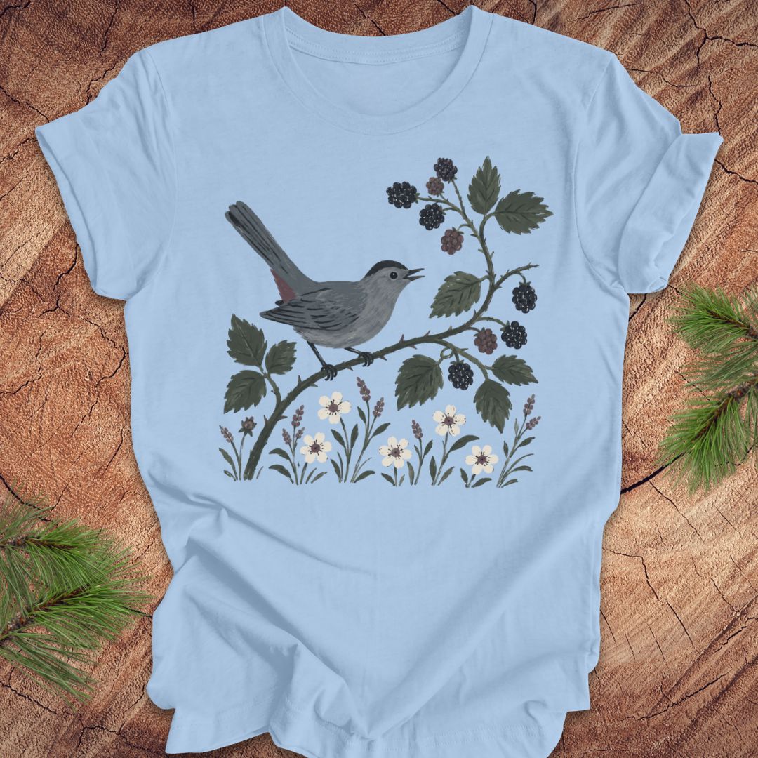 Light blue t-shirt with a catbird and branch design on a wooden surface