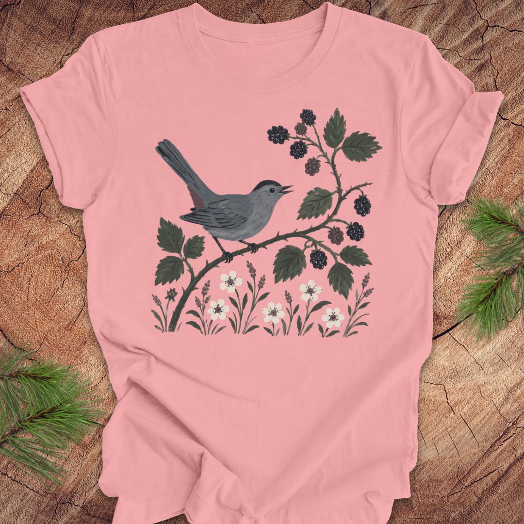 Pink t-shirt with a catbird and floral design on a wooden surface