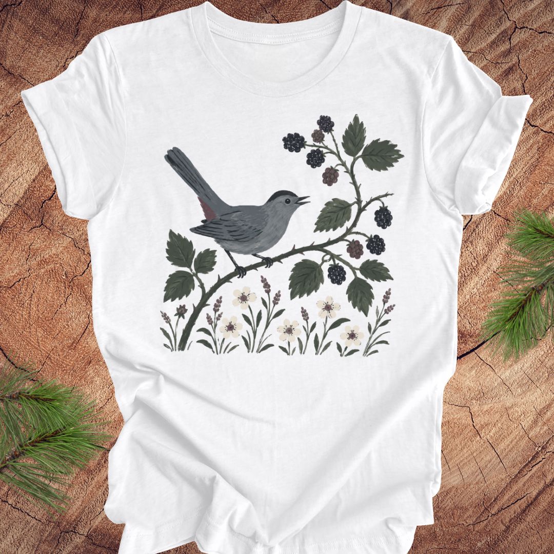 White t-shirt with a catbird and floral design on a wooden surface