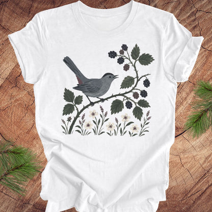 White t-shirt with a catbird and floral design on a wooden surface