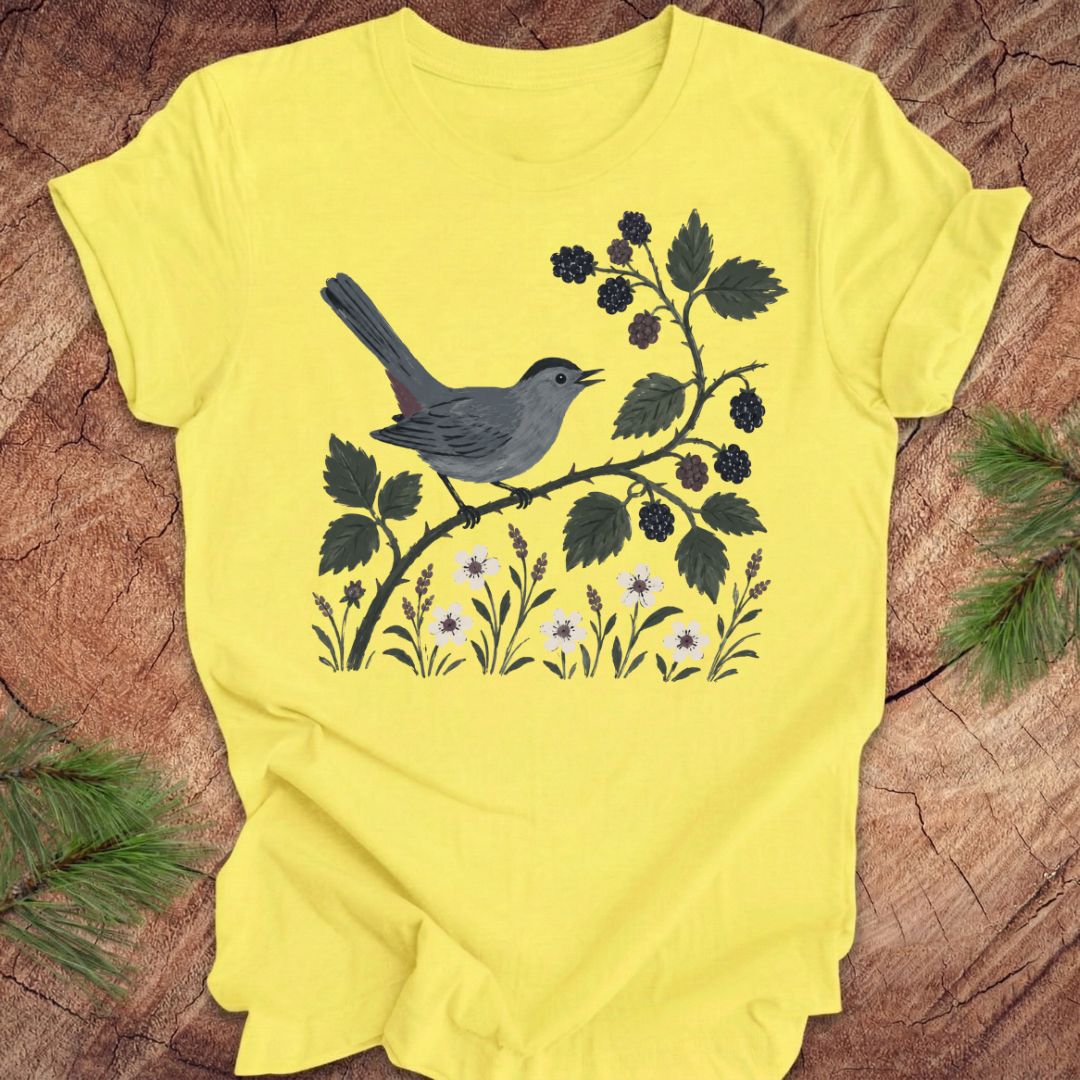 Yellow t-shirt with a catbird and branch design on a wooden surface
