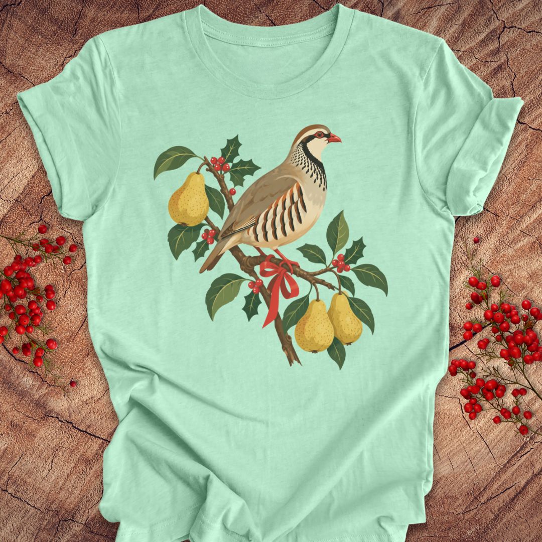 Heather mint green t-shirt with a partridge perched on a branch with golden pears, holly, red berries, and a red ribbon, laid flat on a rustic wood background with red winter berries.